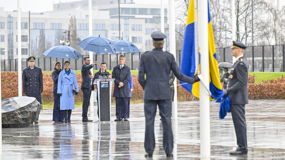 Statsminister Ulf Kristersson håller tal, omgiven av ÖB Micael Bydén, kronprinsessan Victoria och Natochefen Jens Stoltenberg. I förgrunden gör sig sergeant Jonatan Karlstrand och överstelöjtnant André Saeden redo att hissa den svenska flaggan.