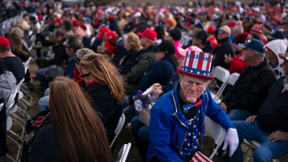 En Trump-anhängare utklädd till Uncle Sam lyssnar medan olika talare värmer upp scenen inför ex-presidentens framträdande.